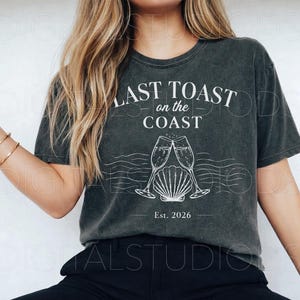 May include: Dark gray t-shirt with the text "LAST TOAST on the COAST" above a graphic of two champagne flutes clinking over a seashell. The text "Est. 2026" is below the graphic. The shirt has a vintage, worn look.