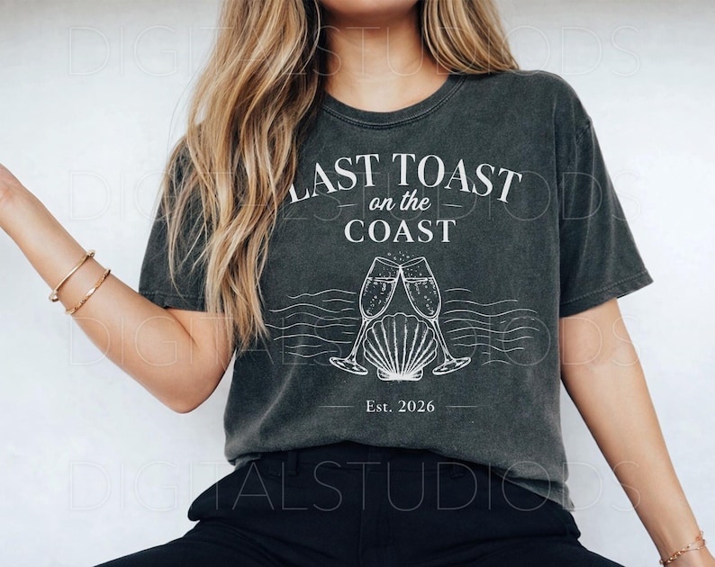 May include: Dark gray t-shirt with the text "LAST TOAST on the COAST" above a graphic of two champagne flutes clinking over a seashell. The text "Est. 2026" is below the graphic. The shirt has a vintage, worn look.
