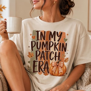 May include: A cream-colored short-sleeve t-shirt featuring the text "IN MY PUMPKIN PATCH ERA" in dark brown letters. The design includes a graphic of an orange pumpkin with a plaid bow, surrounded by autumn leaves, small hearts, and stars.