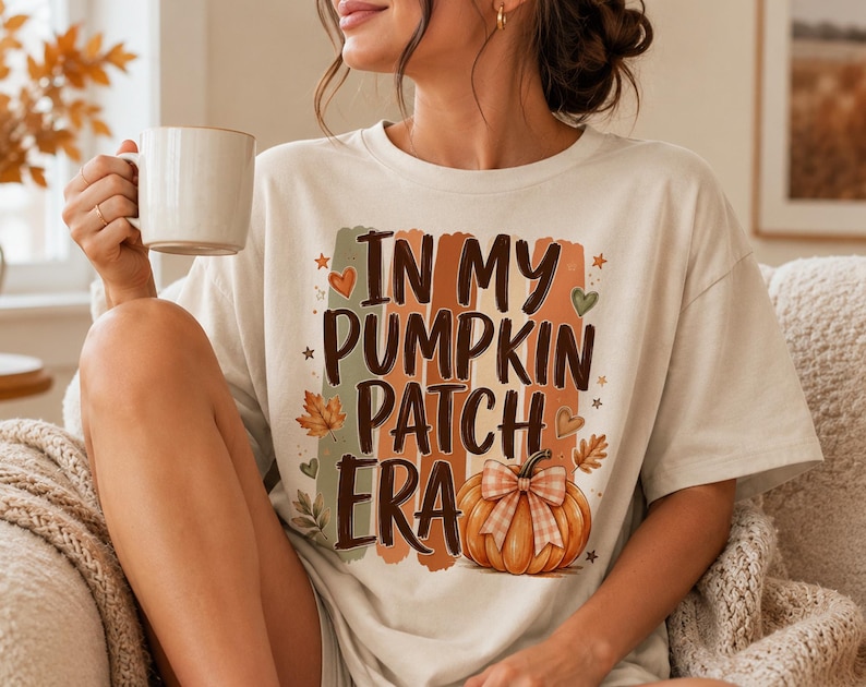 May include: A cream-colored short-sleeve t-shirt featuring the text "IN MY PUMPKIN PATCH ERA" in dark brown letters. The design includes a graphic of an orange pumpkin with a plaid bow, surrounded by autumn leaves, small hearts, and stars.
