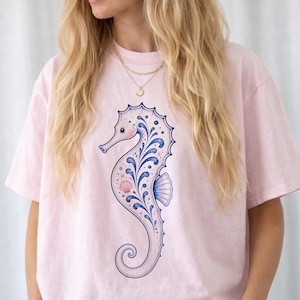 May include: A light pink t-shirt featuring a detailed seahorse design. The seahorse is primarily light pink with blue and white decorative elements. The design includes a seashell and a curved tail.