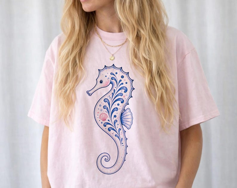 May include: A light pink t-shirt featuring a detailed seahorse design. The seahorse is primarily light pink with blue and white decorative elements. The design includes a seashell and a curved tail.
