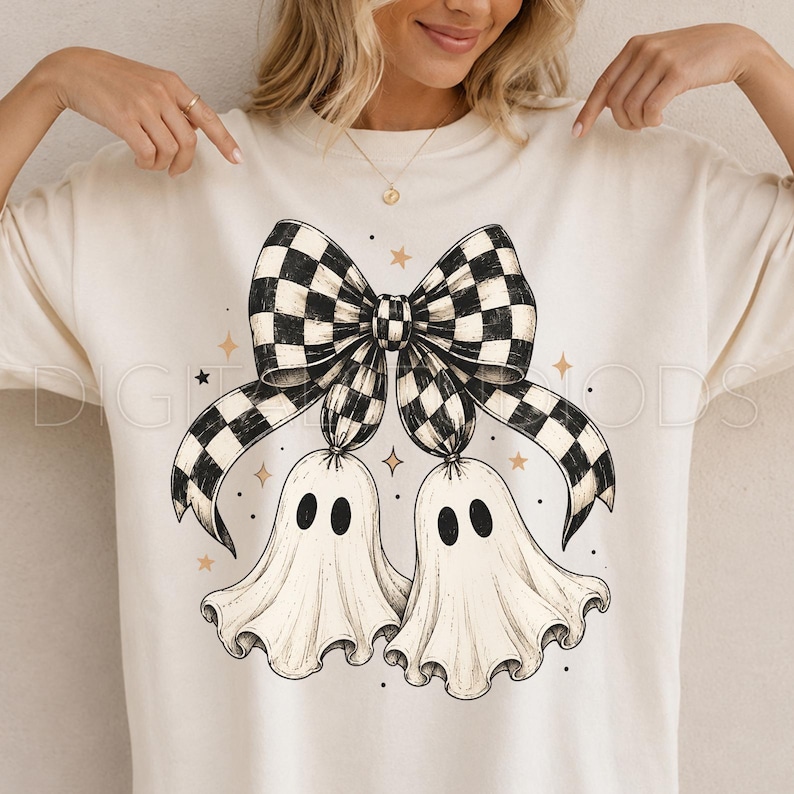 Op de afbeelding: Een cr&egrave;mekleurig t-shirt met een grafische print van twee witte geesten met zwarte ovale ogen die aan een grote zwart-wit geblokte strik hangen. Het ontwerp is versierd met kleine gouden sterretjes en zwarte stippen rondom de illustratie.