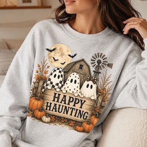 Op de afbeelding: Een lichtgrijs sweatshirt met ronde hals en een grafische print van drie geesten achter een houten bord met de tekst HAPPY HAUNTING. Het ontwerp bevat een schuur, een windmolen, een volle maan met vleermuizen, herfstbladeren en oranje pompoenen.
