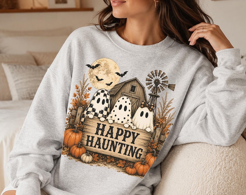 Op de afbeelding: Een lichtgrijs sweatshirt met ronde hals en een grafische print van drie geesten achter een houten bord met de tekst HAPPY HAUNTING. Het ontwerp bevat een schuur, een windmolen, een volle maan met vleermuizen, herfstbladeren en oranje pompoenen.