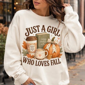 May include: A cream-colored crewneck sweatshirt featuring a graphic print with the text JUST A GIRL WHO LOVES FALL. The design includes a coffee cup labeled PUMPKIN SPICE SEASON, a green book titled COZY STORIES, a mug, and an orange pumpkin.