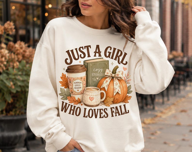 May include: A cream-colored crewneck sweatshirt featuring a graphic print with the text JUST A GIRL WHO LOVES FALL. The design includes a coffee cup labeled PUMPKIN SPICE SEASON, a green book titled COZY STORIES, a mug, and an orange pumpkin.