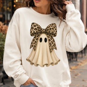 Puede incluir: Una sudadera de manga larga color crema con un gr&aacute;fico central de un fantasma con un gran lazo de estampado de leopardo. El fantasma tiene dos ojos ovalados negros y un dise&ntilde;o de cuerpo drapeado y plisado, centrado en la parte delantera.