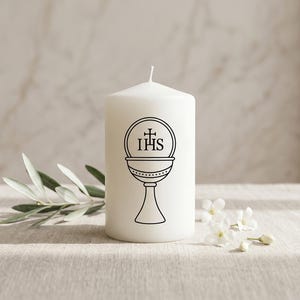 May include: A white pillar candle featuring a black chalice graphic and the letters IHS. The candle is placed on a light surface, accompanied by olive leaves and small white flowers.