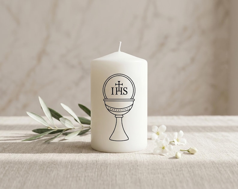 May include: A white pillar candle featuring a black chalice graphic and the letters IHS. The candle is placed on a light surface, accompanied by olive leaves and small white flowers.