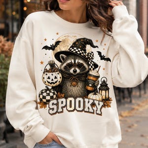 Op de afbeelding: Een cr&egrave;mekleurige sweater met ronde hals en een afbeelding van een wasbeer met een heksenhoed en hartvormige bril. De wasbeer houdt een pompoenemmer en een drankje vast. Het woord SPOOKY staat eronder gedrukt in grote letters.