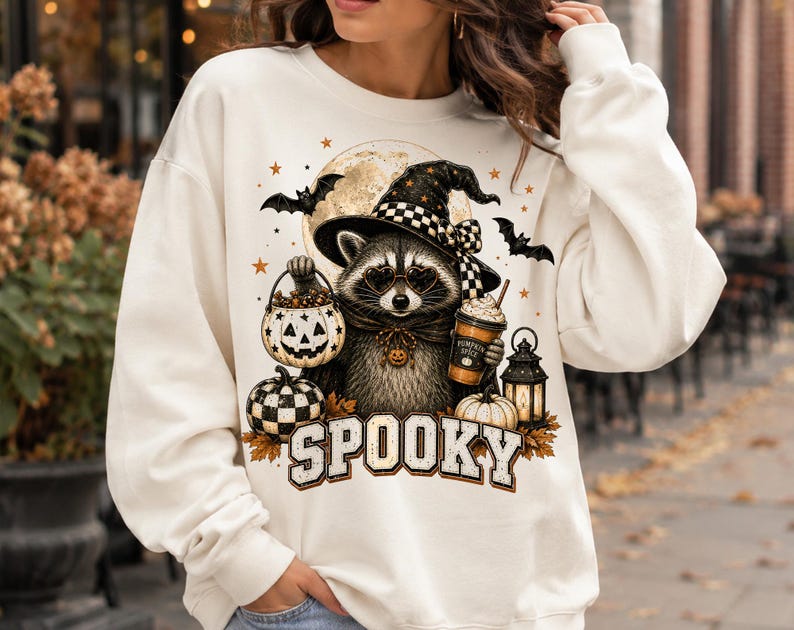 Op de afbeelding: Een cr&egrave;mekleurige sweater met ronde hals en een afbeelding van een wasbeer met een heksenhoed en hartvormige bril. De wasbeer houdt een pompoenemmer en een drankje vast. Het woord SPOOKY staat eronder gedrukt in grote letters.