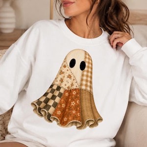 Puede incluir: Una sudadera blanca de cuello redondo con un gr&aacute;fico central de un fantasma compuesto por varios patrones de tela. El dise&ntilde;o del fantasma incluye secciones de cuadros vichy en marr&oacute;n y crema, estampados florales y un patr&oacute;n de cuadros, todo con dos ojos ovalados negros.