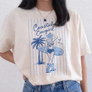 May include: Cream-colored t-shirt featuring a blue graphic of a woman with a surfboard, palm tree, and the text "Coastal Cowgirl." The design includes vertical blue stripes and a cowgirl in a hat and boots.