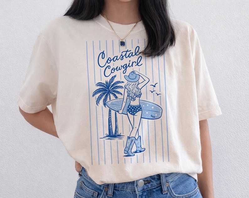 May include: Cream-colored t-shirt featuring a blue graphic of a woman with a surfboard, palm tree, and the text "Coastal Cowgirl." The design includes vertical blue stripes and a cowgirl in a hat and boots.