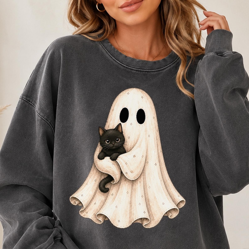Puede incluir: Una sudadera de cuello redondo gris carb&oacute;n con un estampado gr&aacute;fico de un fantasma color crema envuelto en una s&aacute;bana, sosteniendo un peque&ntilde;o gato negro de ojos grandes. El fantasma tiene dos ojos ovalados negros y aspecto fluido.