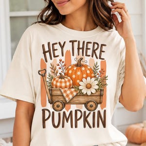 May include: A cream-colored t-shirt featuring a graphic of a wooden wagon filled with two pumpkins, a white daisy, and foliage. The text "HEY THERE PUMPKIN" is printed in brown above and below the wagon illustration, which includes orange brushstrokes.
