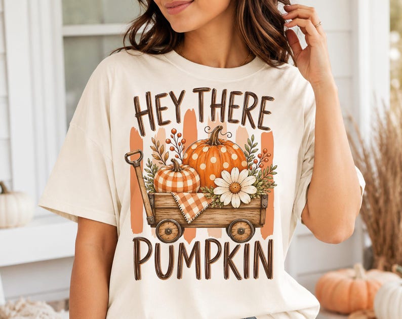 May include: A cream-colored t-shirt featuring a graphic of a wooden wagon filled with two pumpkins, a white daisy, and foliage. The text "HEY THERE PUMPKIN" is printed in brown above and below the wagon illustration, which includes orange brushstrokes.
