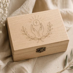 May include: A light-coloured wooden box with a hinged lid and a metal clasp. The top of the box features an engraved design of hands holding a cross and a circle, surrounded by a laurel wreath. The box is resting on a beige fabric.