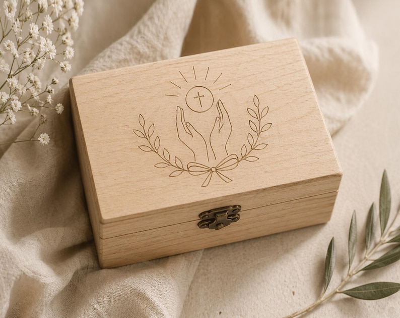 May include: A light-coloured wooden box with a hinged lid and a metal clasp. The top of the box features an engraved design of hands holding a cross and a circle, surrounded by a laurel wreath. The box is resting on a beige fabric.