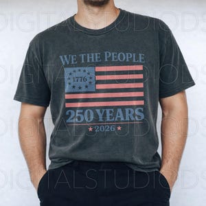 May include: Dark gray t-shirt featuring the text "WE THE PEOPLE" above a stylized American flag design with the text "1776" and "250 YEARS 2026". The shirt has a vintage, worn look.