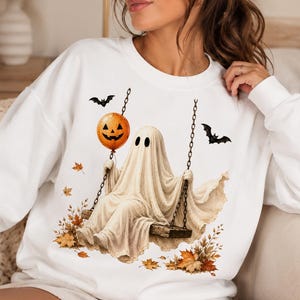 Op de afbeelding: Een wit sweatshirt met lange mouwen en een grafische print van een geest die op een houten schommel zit. De geest houdt een oranje pompoenballon vast. Het ontwerp bevat twee zwarte vleermuizen en verspreide herfstbladeren in oranje en bruin.