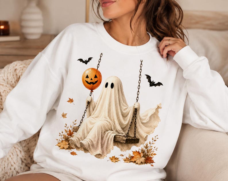 Op de afbeelding: Een wit sweatshirt met lange mouwen en een grafische print van een geest die op een houten schommel zit. De geest houdt een oranje pompoenballon vast. Het ontwerp bevat twee zwarte vleermuizen en verspreide herfstbladeren in oranje en bruin.