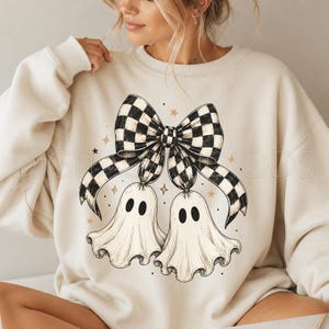 Op de afbeelding: Een cr&egrave;mekleurige sweater met ronde hals en een grafische print van twee witte geesten die aan een grote zwart-wit geblokte strik hangen. Het ontwerp bevat kleine goudkleurige steraccenten verspreid rond de centrale illustratie.