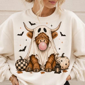 May include: A cream-colored crewneck sweatshirt featuring a graphic of a brown Highland cow wearing a white ghost sheet, blowing a pink bubblegum bubble. The design includes black bats, orange stars, a checkered pumpkin, a plain pumpkin, and a lantern.