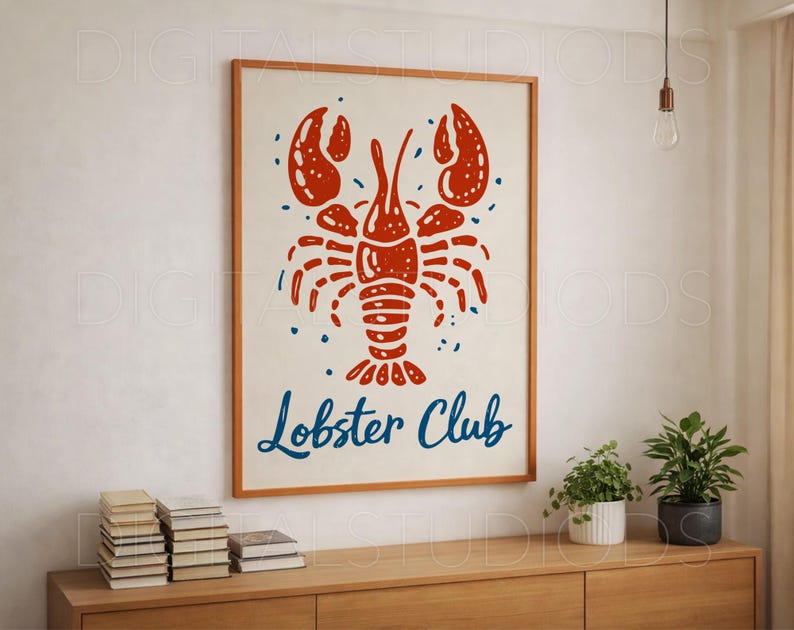 May include: A framed print featuring a red lobster illustration with the words "Lobster Club" in blue script. The artwork is set against a cream background and framed in a light brown wooden frame. The print is displayed on a wooden cabinet.