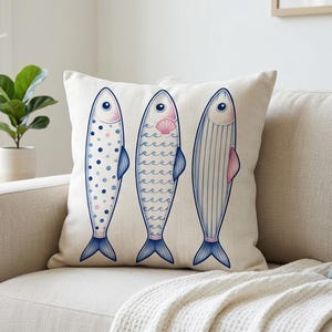 May include: A decorative throw pillow featuring three stylized fish illustrations. Each fish has a different pattern: polka dots, waves, and vertical lines. The fish are in shades of blue, pink, and white on a cream-colored background. The pillow is placed on a beige sofa.