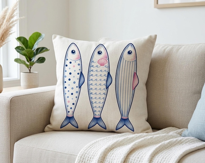 May include: A decorative throw pillow featuring three stylized fish illustrations. Each fish has a different pattern: polka dots, waves, and vertical lines. The fish are in shades of blue, pink, and white on a cream-colored background. The pillow is placed on a beige sofa.