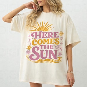 May include: Cream-colored oversized t-shirt with the text "HERE COMES THE SUN" in pink and yellow. The design includes a sun, flowers, and waves. The shirt has short sleeves and a relaxed fit.