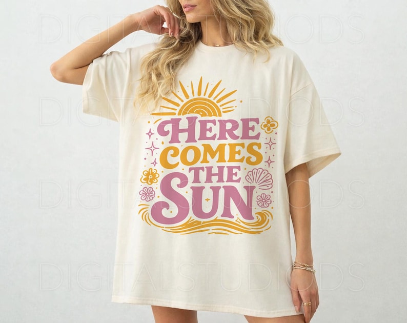 May include: Cream-colored oversized t-shirt with the text "HERE COMES THE SUN" in pink and yellow. The design includes a sun, flowers, and waves. The shirt has short sleeves and a relaxed fit.