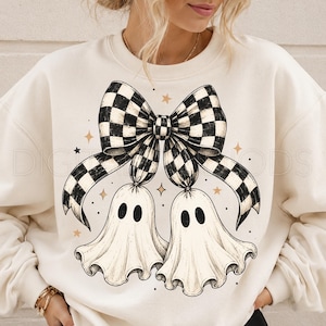 Op de afbeelding: Een cr&egrave;mekleurig sweatshirt met ronde hals en een grote grafische print van twee witte geesten die aan een zwart-wit geblokte strik hangen. Het ontwerp bevat kleine gouden steraccenten en zwarte stippen rond de illustratie.