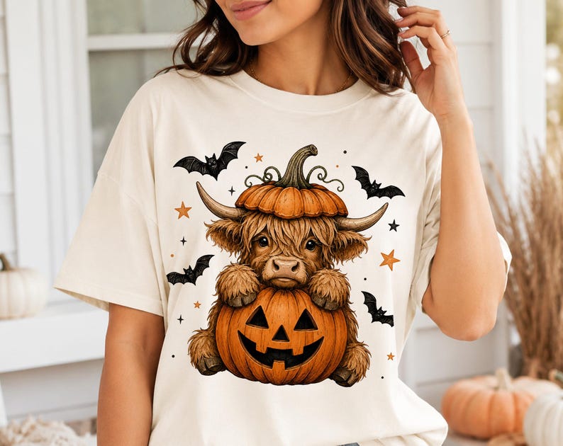 Op de afbeelding: Een cr&egrave;mekleurig t-shirt met een afbeelding van een pluizige bruine Schotse hooglander met een pompoen op zijn kop, zittend achter een uitgesneden pompoen. Het ontwerp bevat zwarte vleermuizen en oranje sterren rondom de centrale figuur.