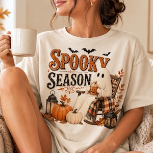 Op de afbeelding: Een cr&egrave;mekleurig t-shirt met een grafische print met de tekst "SPOOKY SEASON" in oranje en zwart. Het ontwerp bevat een geest in een schommelstoel, pompoenen, lantaarns, herfstbladeren en vleermuizen. De geest houdt een kleine mok vast.