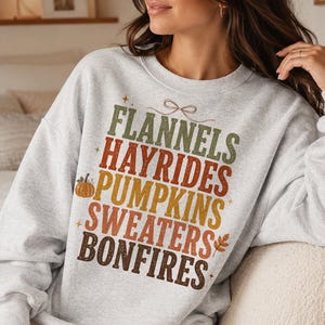 May include: A light gray crewneck sweatshirt featuring a multi-colored graphic print with the stacked text: FLANNELS, HAYRIDES, PUMPKINS, SWEATERS, BONFIRES. The design includes a small orange pumpkin, a brown leaf, and a thin line-art bow graphic.
