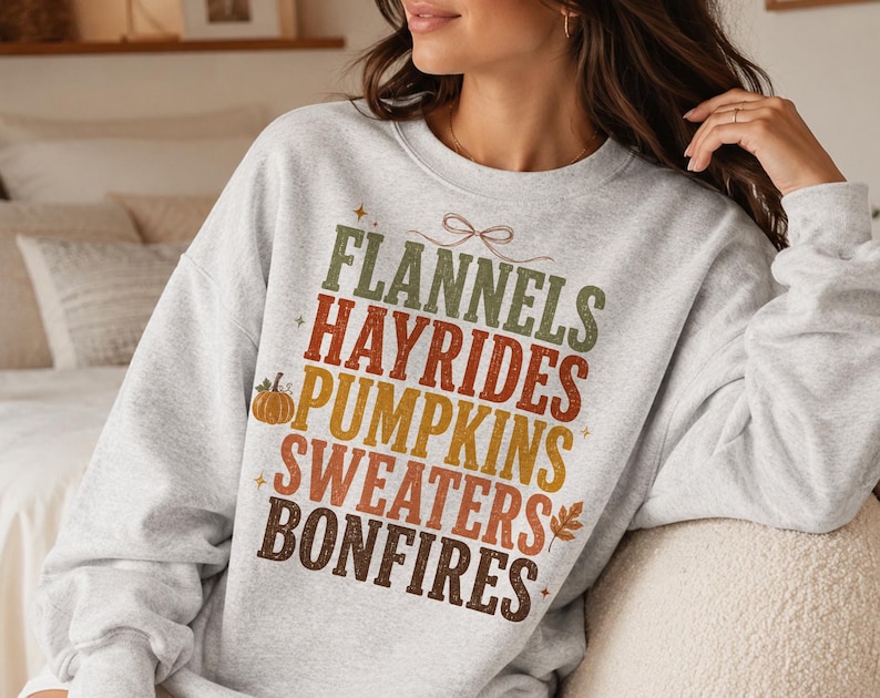 May include: A light gray crewneck sweatshirt featuring a multi-colored graphic print with the stacked text: FLANNELS, HAYRIDES, PUMPKINS, SWEATERS, BONFIRES. The design includes a small orange pumpkin, a brown leaf, and a thin line-art bow graphic.