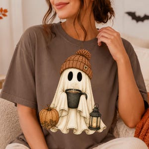 Puede incluir: Una camiseta de manga corta de color marr&oacute;n oscuro con el gr&aacute;fico de un fantasma blanco que lleva un gorro de punto marr&oacute;n, sostiene una taza de caf&eacute; negra y est&aacute; junto a una calabaza naranja y un farol negro con luz interior.