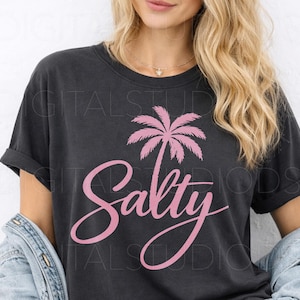 May include: Dark gray t-shirt with a pink palm tree graphic and the word "Salty" in a cursive font. The shirt has rolled-up sleeves and a relaxed fit. The design is centered on the front of the shirt.