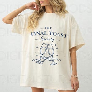 May include: Cream-colored t-shirt with the text "THE FINAL TOAST Society" in navy blue, along with an illustration of two champagne flutes clinking. The design includes stars and a bow. The shirt has short sleeves.