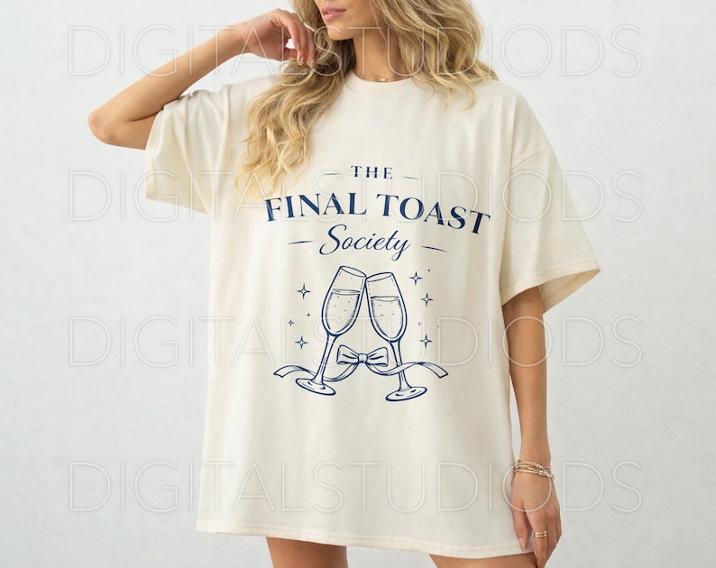 May include: Cream-colored t-shirt with the text "THE FINAL TOAST Society" in navy blue, along with an illustration of two champagne flutes clinking. The design includes stars and a bow. The shirt has short sleeves.