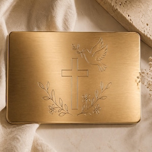 May include: A rectangular, gold-coloured metal plaque featuring an engraved cross, dove, and floral design. The dove carries an olive branch. The plaque is set against a neutral background with soft fabric and decorative elements.