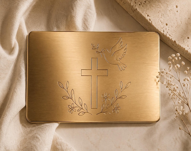 May include: A rectangular, gold-coloured metal plaque featuring an engraved cross, dove, and floral design. The dove carries an olive branch. The plaque is set against a neutral background with soft fabric and decorative elements.