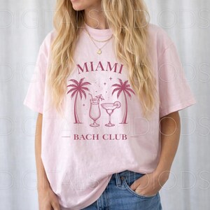 May include: A light pink t-shirt with a graphic design featuring the text "MIAMI BACH CLUB" in a maroon color. The design includes two palm trees and two cocktail glasses. The model is wearing blue jeans.