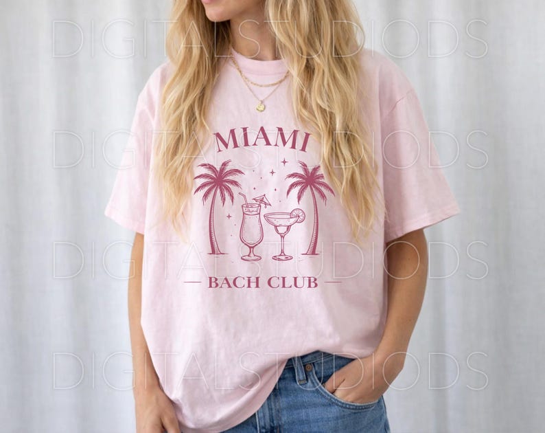 May include: A light pink t-shirt with a graphic design featuring the text "MIAMI BACH CLUB" in a maroon color. The design includes two palm trees and two cocktail glasses. The model is wearing blue jeans.