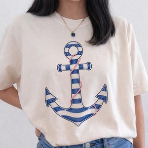 May include: Cream-colored t-shirt featuring a nautical anchor design. The anchor is blue and white striped with a pink rope detail. The shirt has short sleeves and a crew neck, suitable for casual wear.