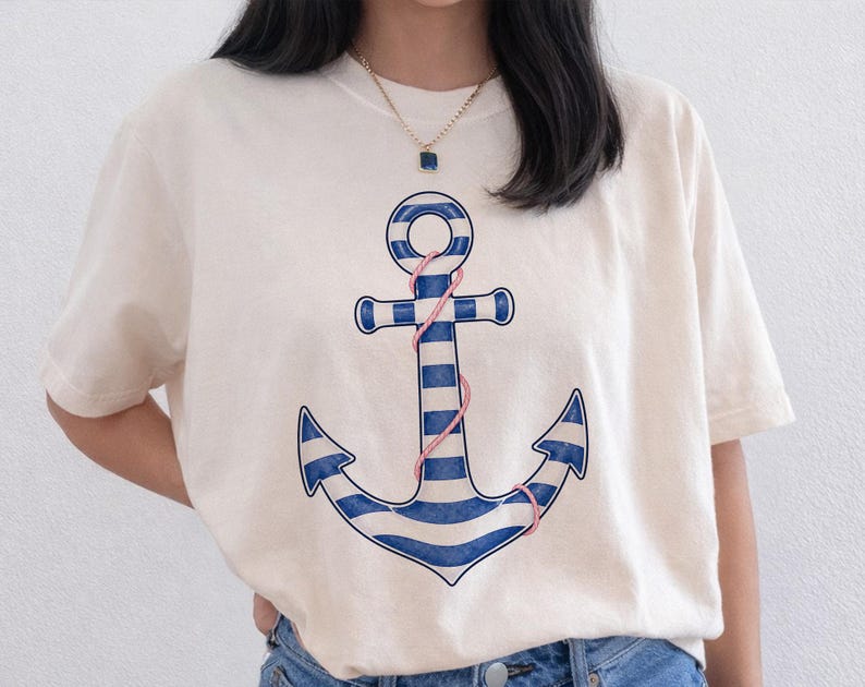 May include: Cream-colored t-shirt featuring a nautical anchor design. The anchor is blue and white striped with a pink rope detail. The shirt has short sleeves and a crew neck, suitable for casual wear.