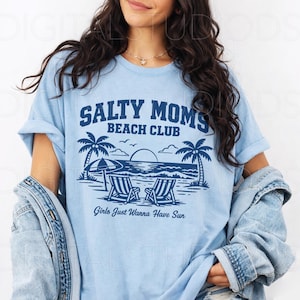 May include: Light blue t-shirt with a graphic that reads "Salty Moms Beach Club" above a beach scene with palm trees, beach chairs, and the text "Girls Just Wanna Have Sun."